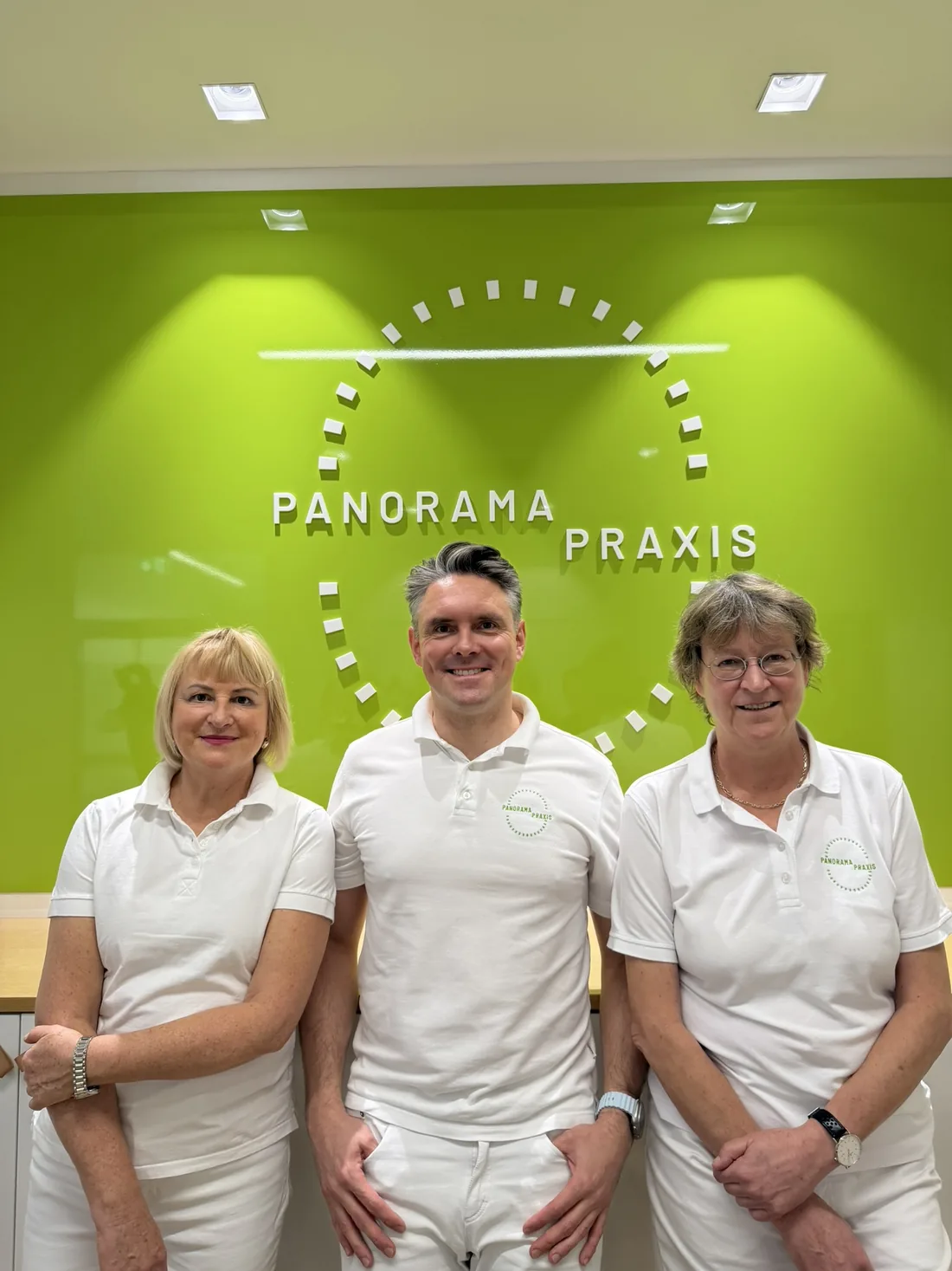 Drei Mitglieder des Teams der Panorama Praxis vor der grünen Logowand